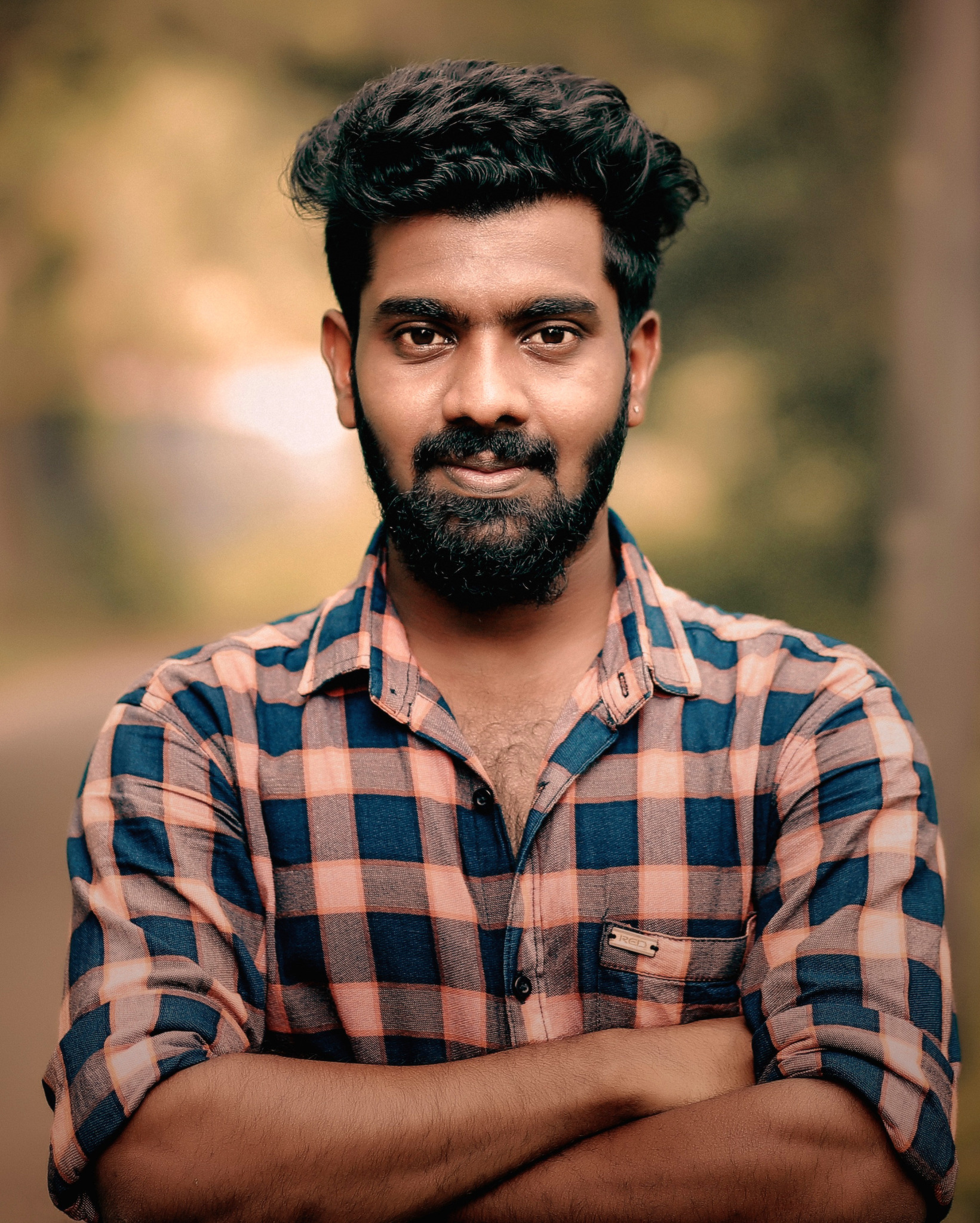 Retrato fotográfico de um homem jovem com barba, de braços cruzados, em ambiente externo.