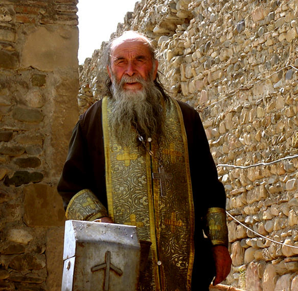 Sacerdote ortodoxo georgiano com estola dourada e cruz, fotografado junto a muros de pedra de local histórico.